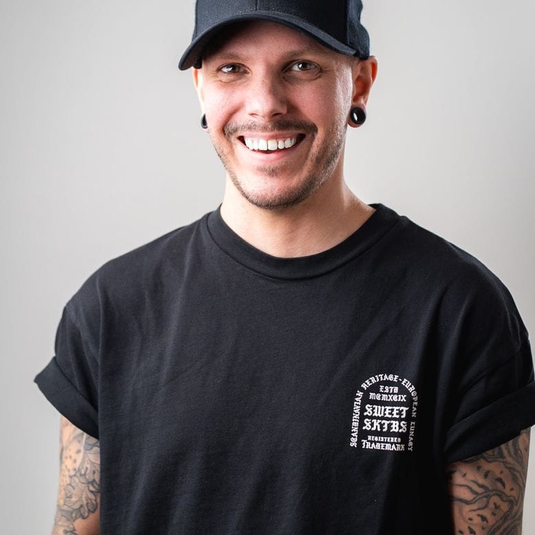 Ung man i svart t-shirt med tatueringar på armarna och en svart keps står mot en grå bakgrund. Han ler och bär stora örhängen.