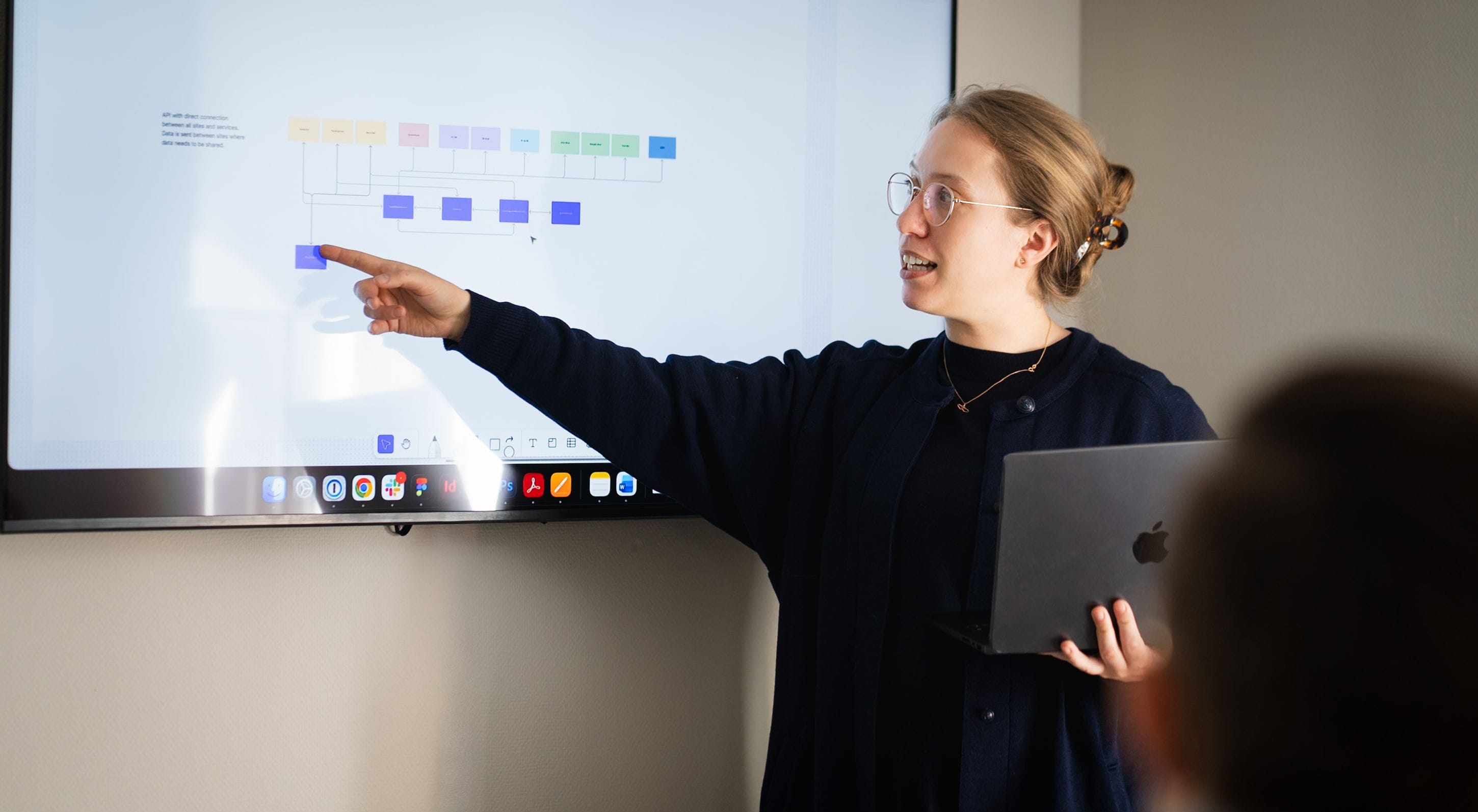 A person with glasses and a laptop points to a chart on a screen in an office.
