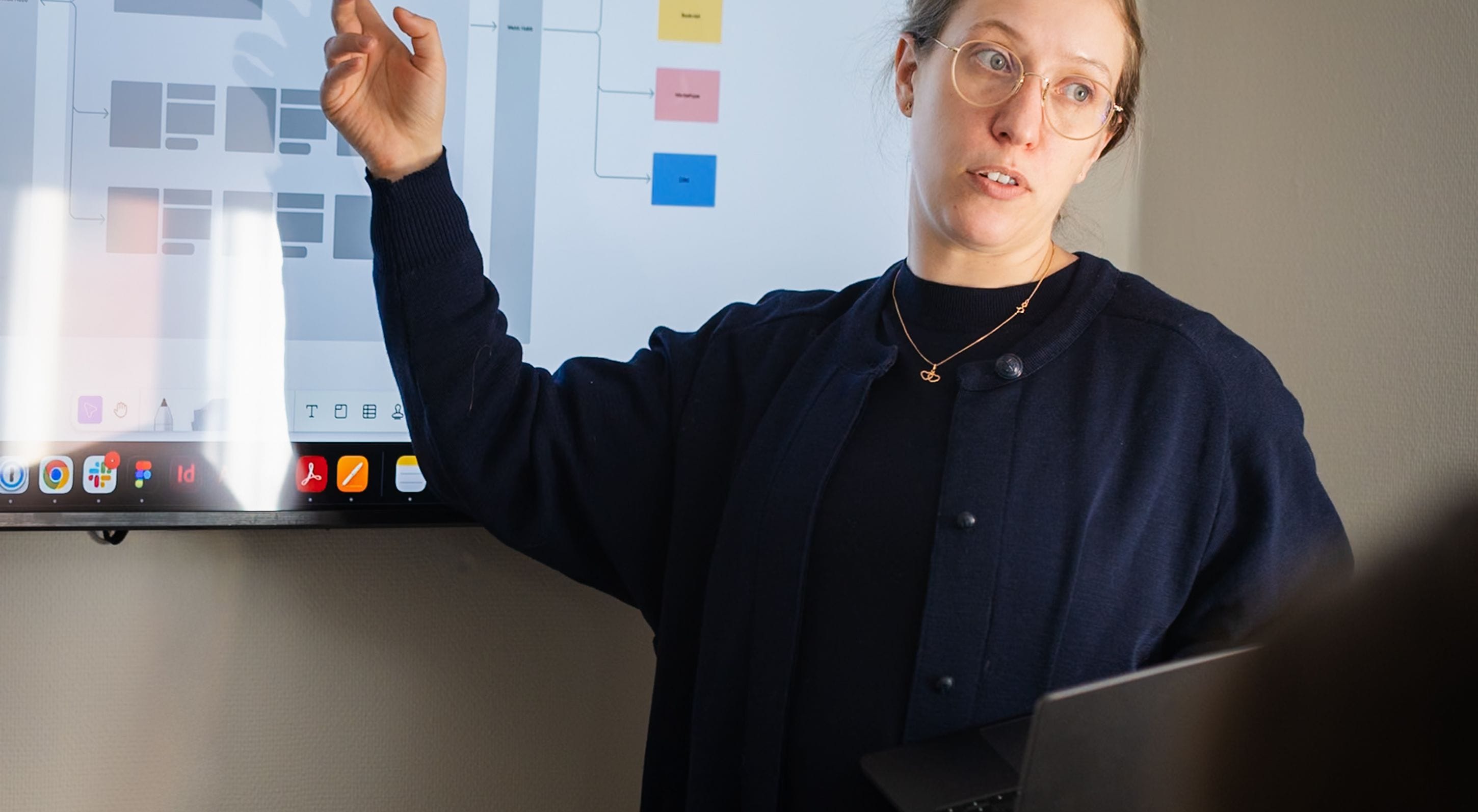 A person gives a presentation in front of a large computer screen displaying a layout diagram. They point at the screen while holding a laptop.