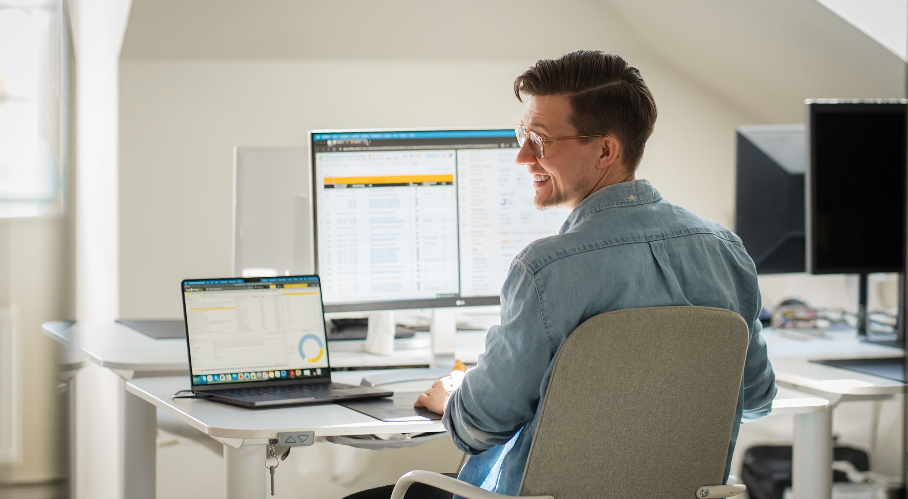 A man in a light blue shirt sits at a desk working on a computer. Several monitors and a laptop are on the desk. He smiles and turns towards the camera in Pigment's office.
