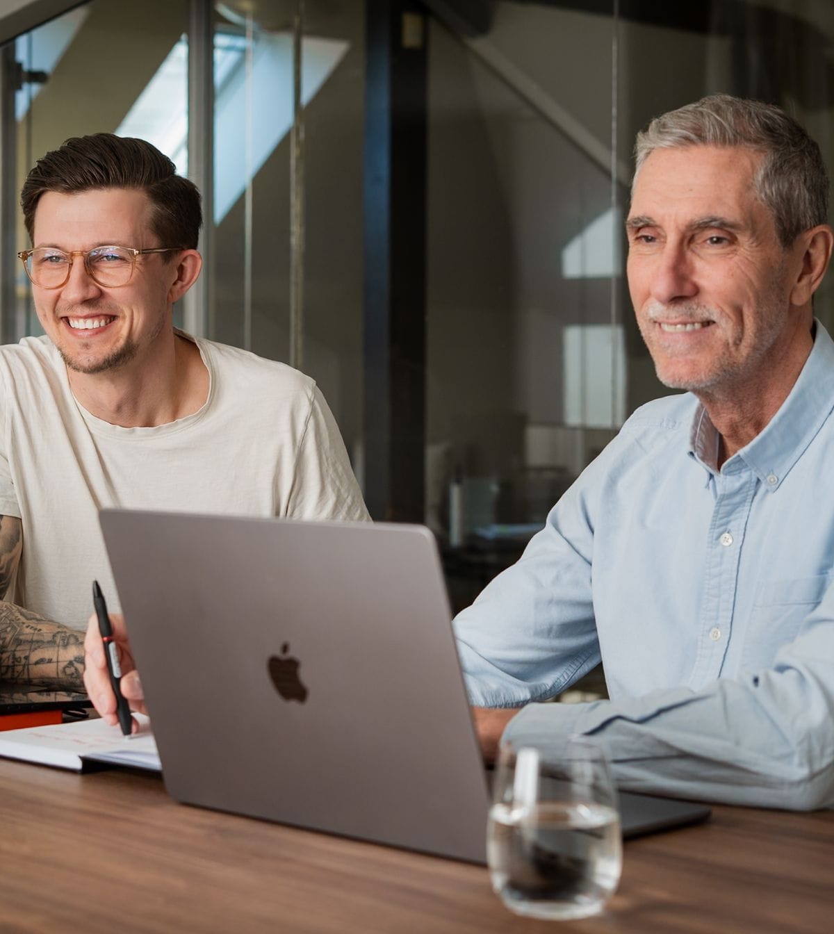 Two men sitting at a table in the Pigment office, each working on a laptop. The younger man wears glasses and has tattoos on one arm. The older man is smiling and wearing a light blue shirt.