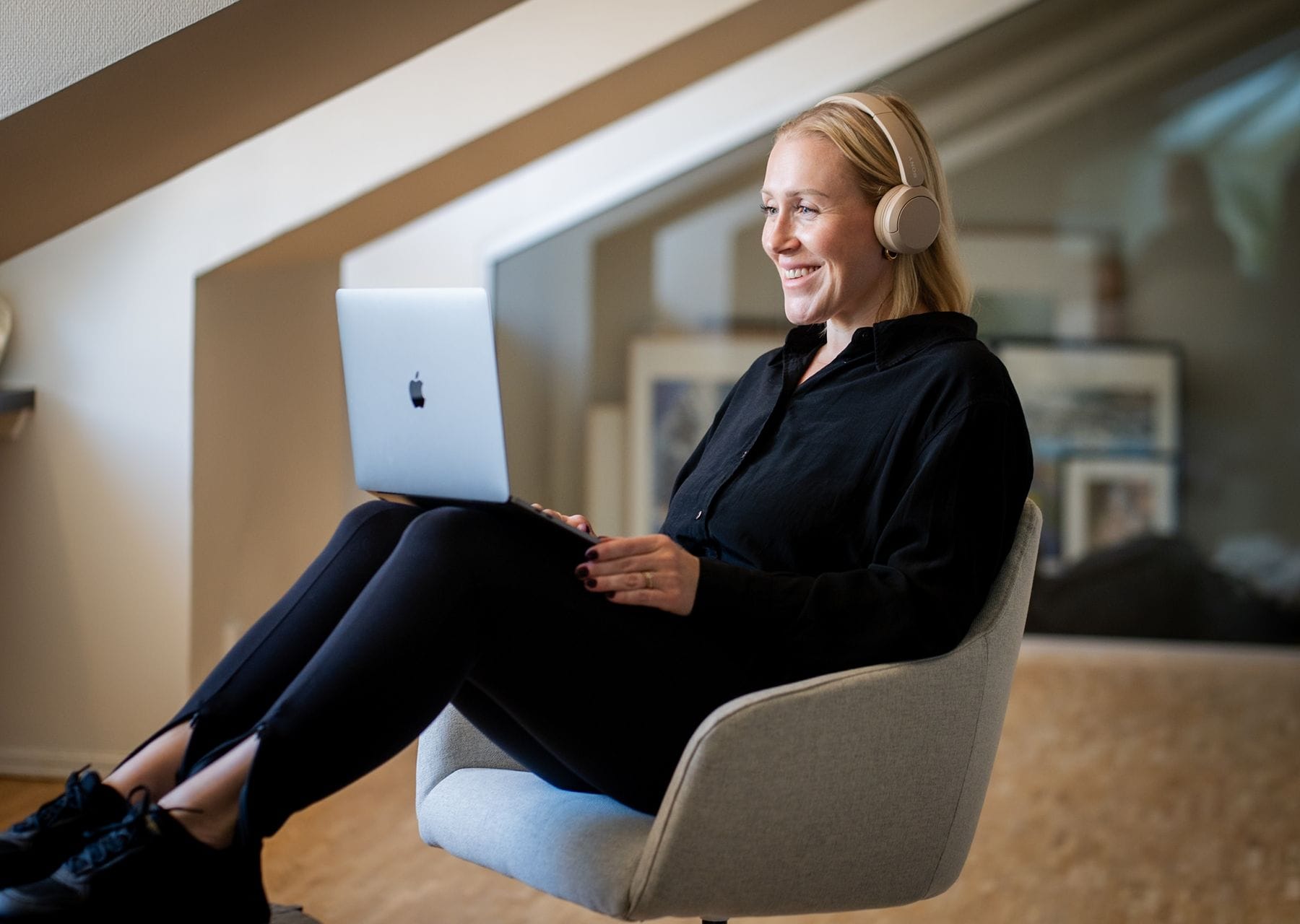 A woman sits comfortably in a grey armchair in a modern office, wearing headphones and smiling as she uses a laptop.
