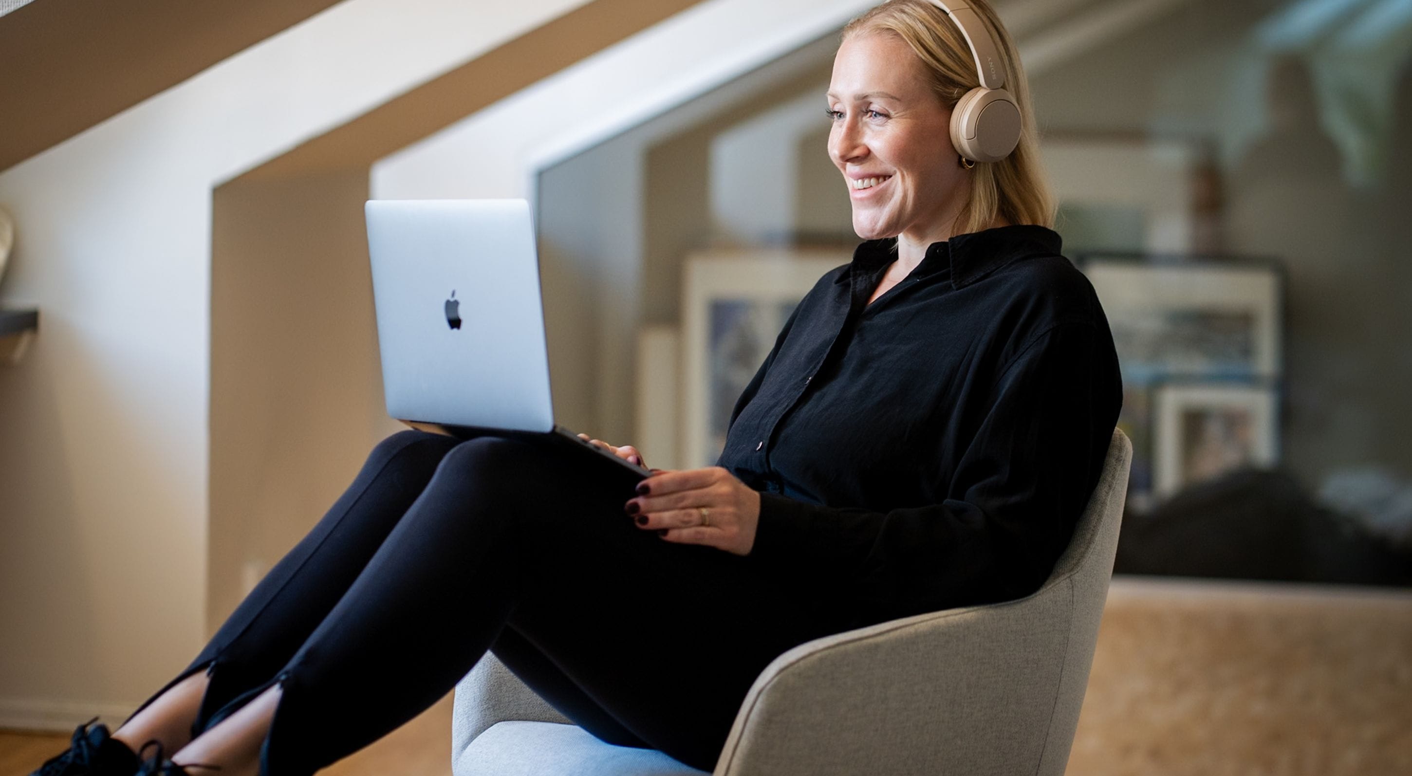 En kvinna sitter avslappnad i en grå fåtölj, bär hörlurar och ler medan hon använder en laptop, i ett modernt kontor.