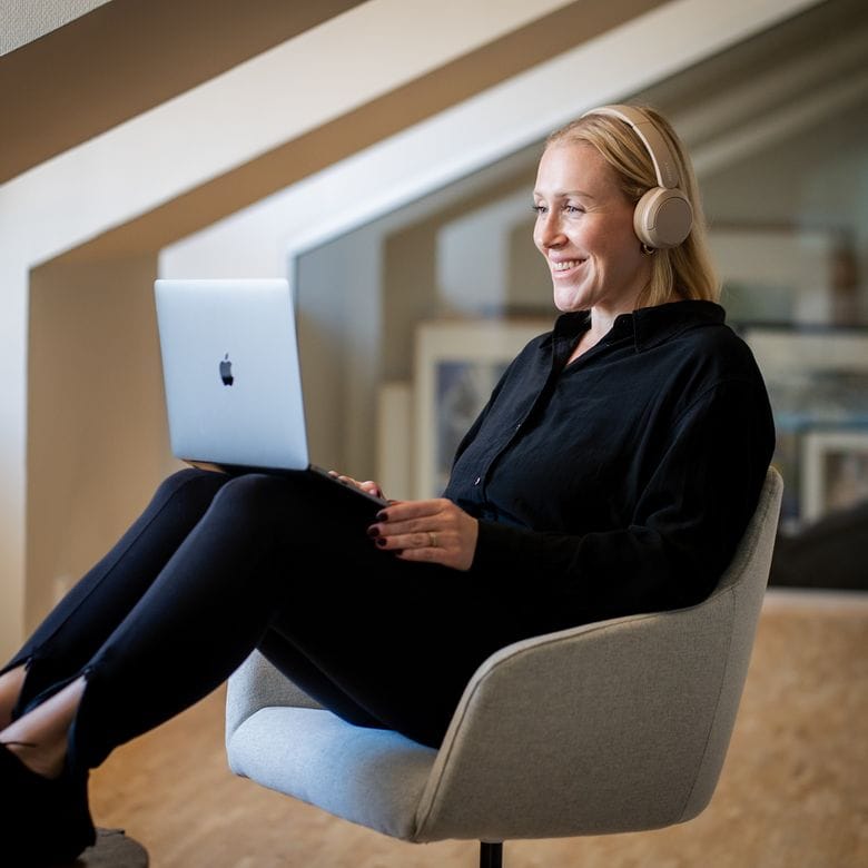 En kvinna sitter avslappnad i en grå fåtölj, bär hörlurar och ler medan hon använder en laptop, i ett modernt kontor.