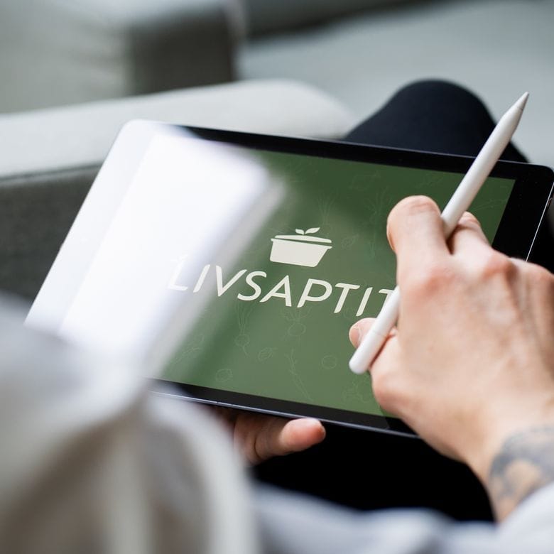 A person holds a tablet with the Livsaptit logo visible on the screen. They use a stylus to interact with the device, with a blurred sofa in the background.