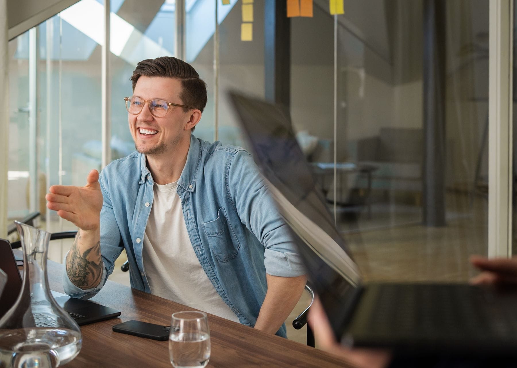 A man wearing glasses and a denim shirt smiles and gestures at a conference table in a modern office.