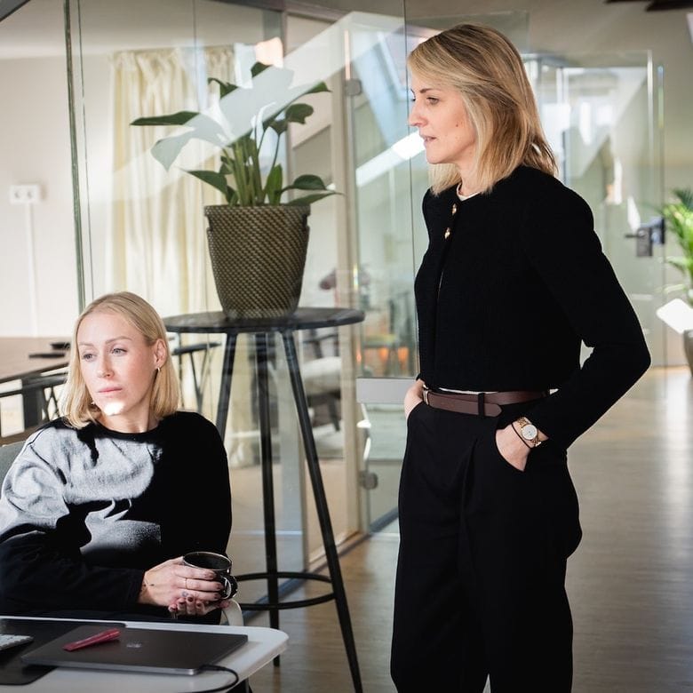 Två kvinnor i ett modernt kontor, en sitter vid en dator med en kaffekopp, den andra står bredvid och tittar på skärmen.