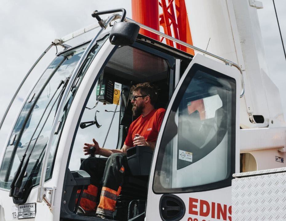 Edins Kranars kranförare sitter i hytten på en stor kran och gestikulerar. Kranen är vit och röd, med klar himmel i bakgrunden.