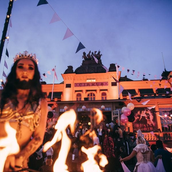 Eldartister och publik framför Cirkus upplysta fasad en sommarkväll, med vimplar över entrén.