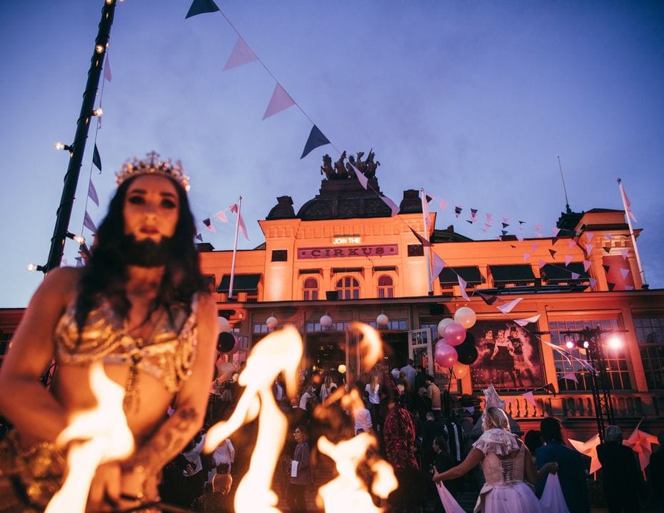 Eldartister och publik framför Cirkus upplysta fasad en sommarkväll, med vimplar över entrén.