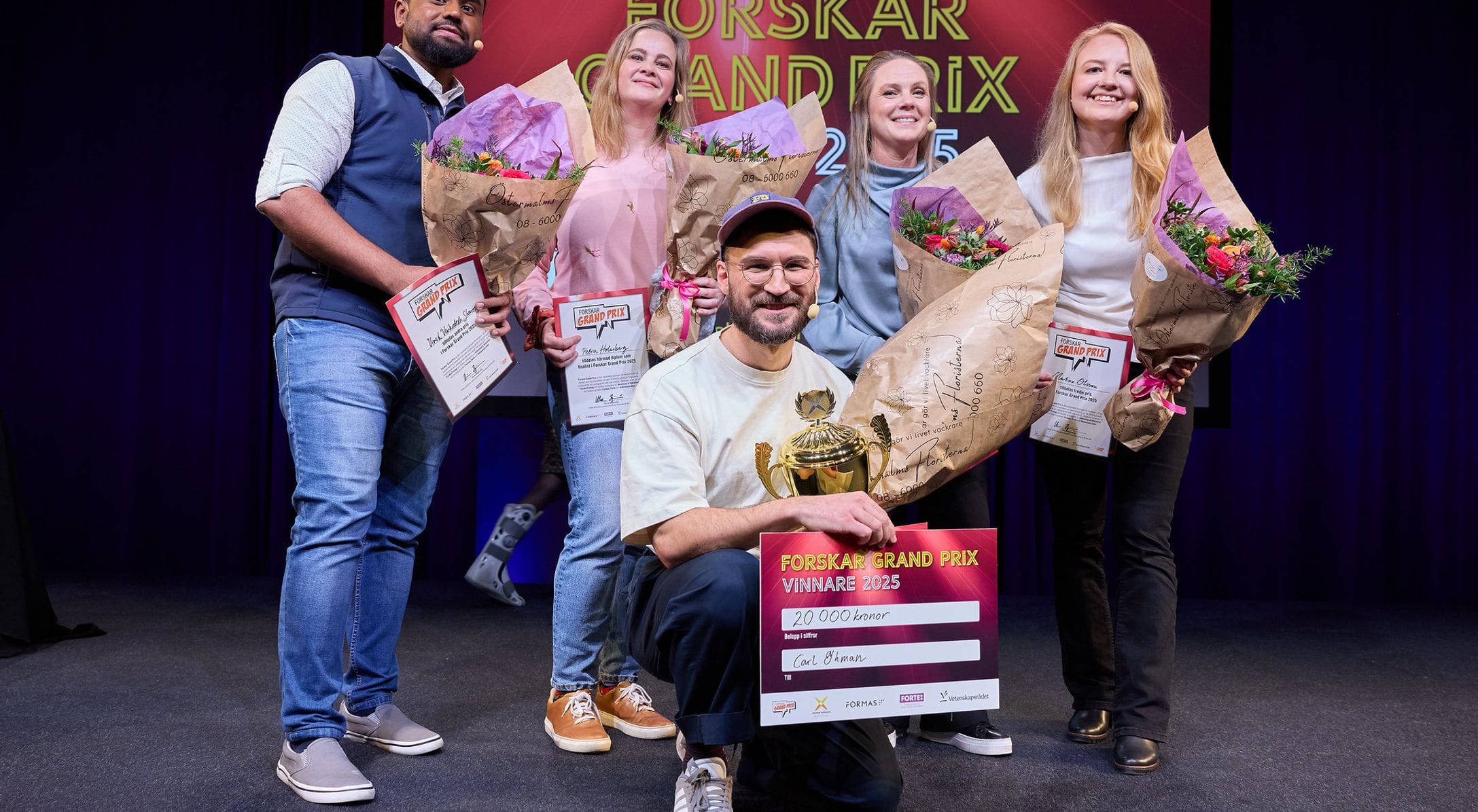 Five people on stage with flowers and a Forskar Grand Prix certificate. The man in the centre holds the certificate and a trophy.