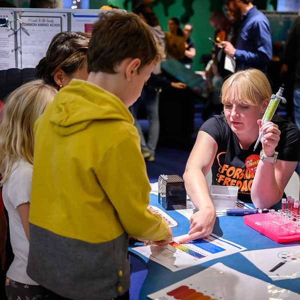 A person at a table during ForskarFredag explains an experiment to children, with test tubes and notes on the table.