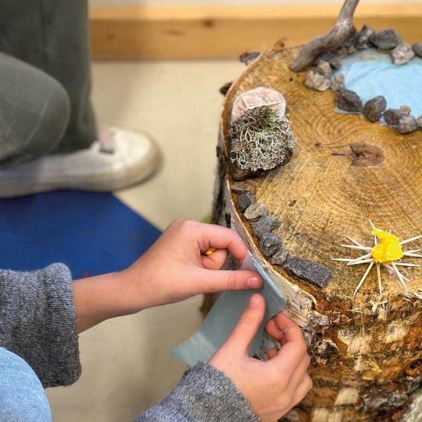 Children decorate a tree stump with small stones, moss and a yellow paper flower as part of a creative project.