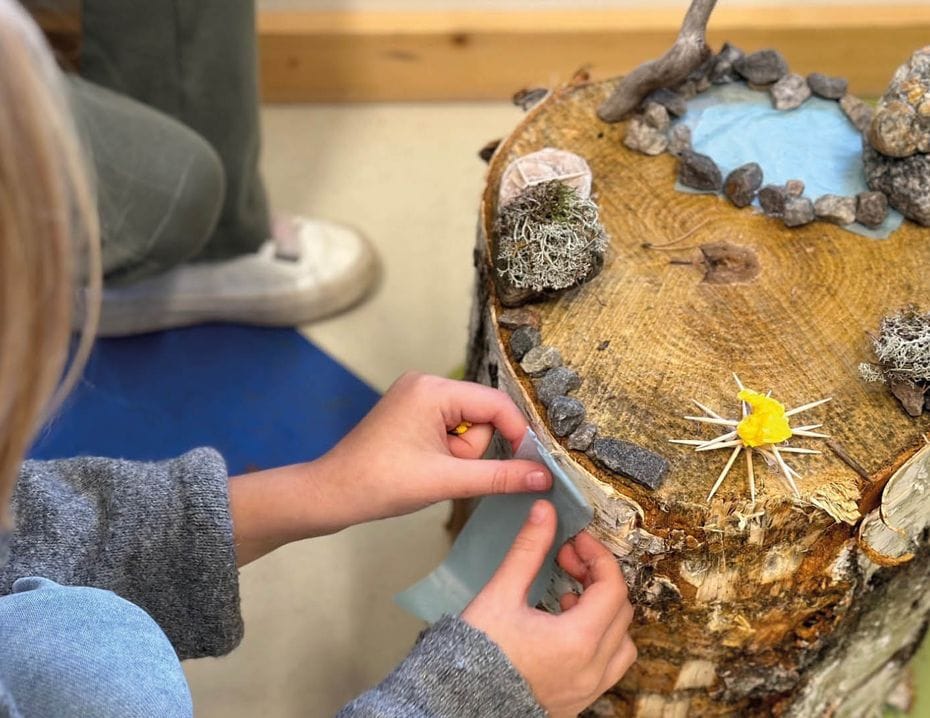 Barn dekorerar en trästubbe med små stenar, mossa och en gul pappersblomma, vilket ser ut som ett kreativt projekt.