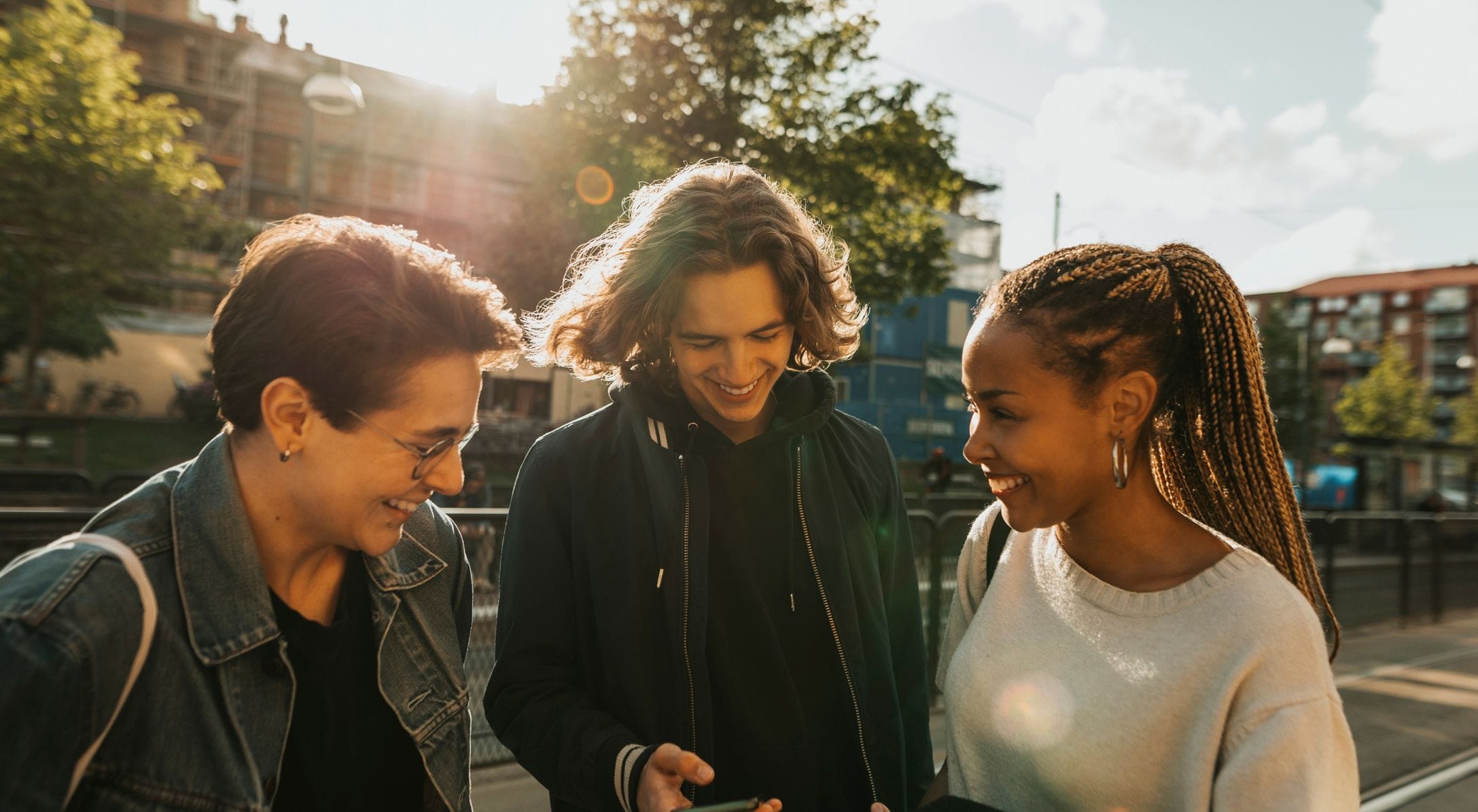 Three people stand outdoors, smiling as they look at a mobile phone. The sun is shining, with trees and buildings in the background.