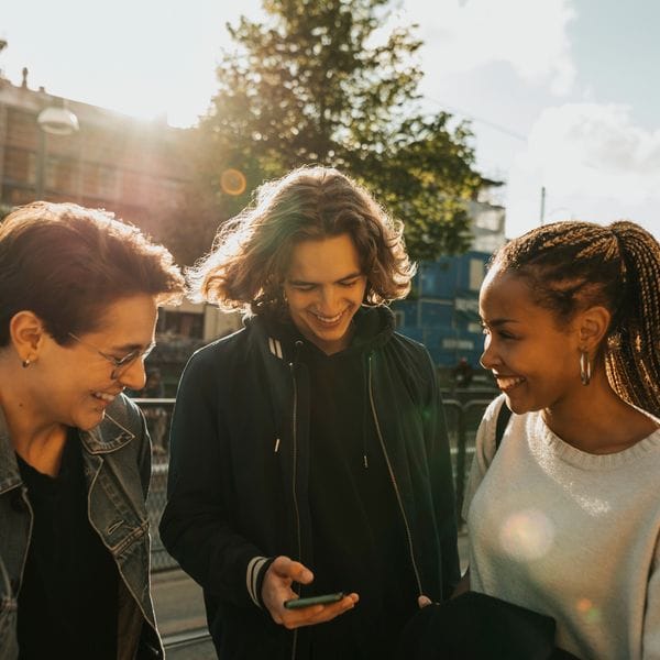 Tre personer står utomhus och ler medan de tittar på en mobiltelefon. Solen skiner, och det finns träd och byggnader i bakgrunden.
