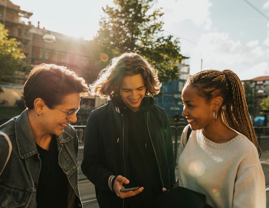 Tre personer står utomhus och ler medan de tittar på en mobiltelefon. Solen skiner, och det finns träd och byggnader i bakgrunden.