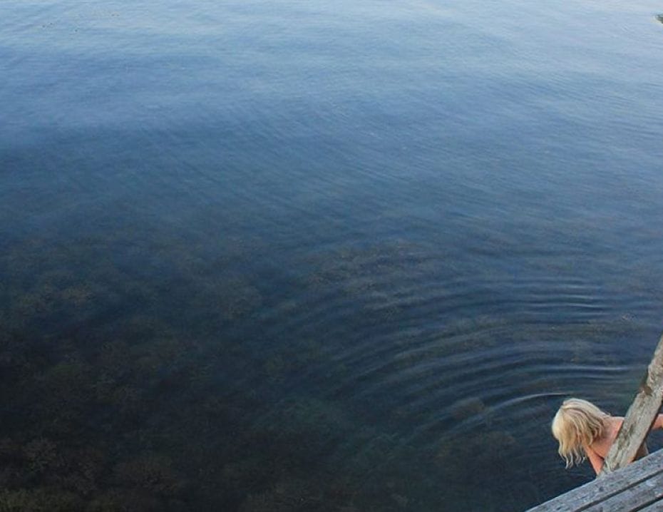 En person går ner i vattnet från en brygga vid en sjö eller hav, omgiven av klippor.