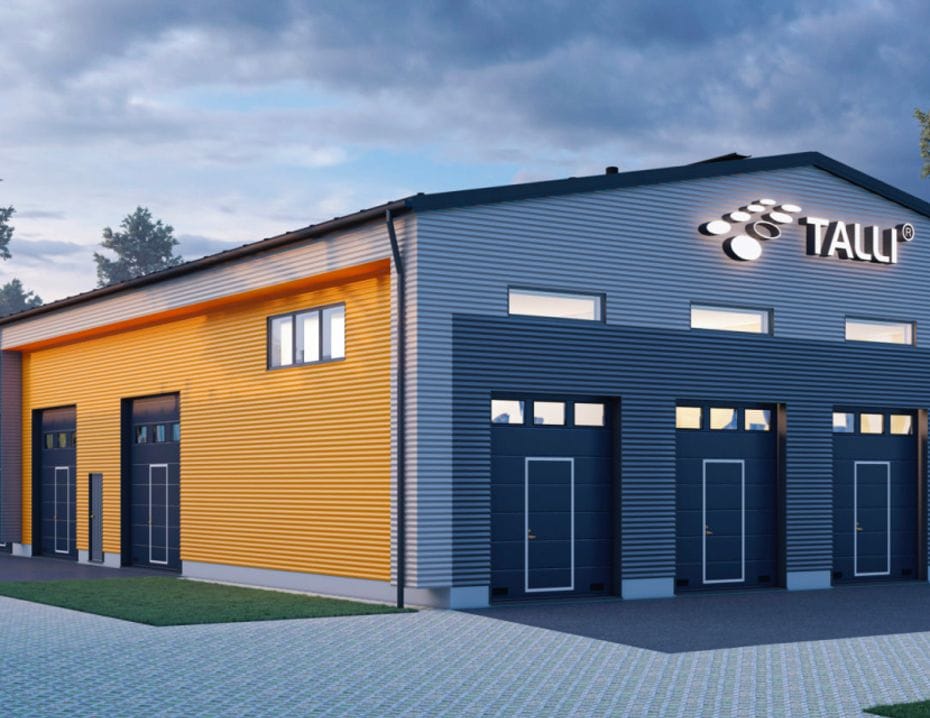 En modern lagerbyggnad med gul och grå fasad, med ett stort upplyst Talli-varumärke på gaveln, omgiven av träd och en stenlagd gång.