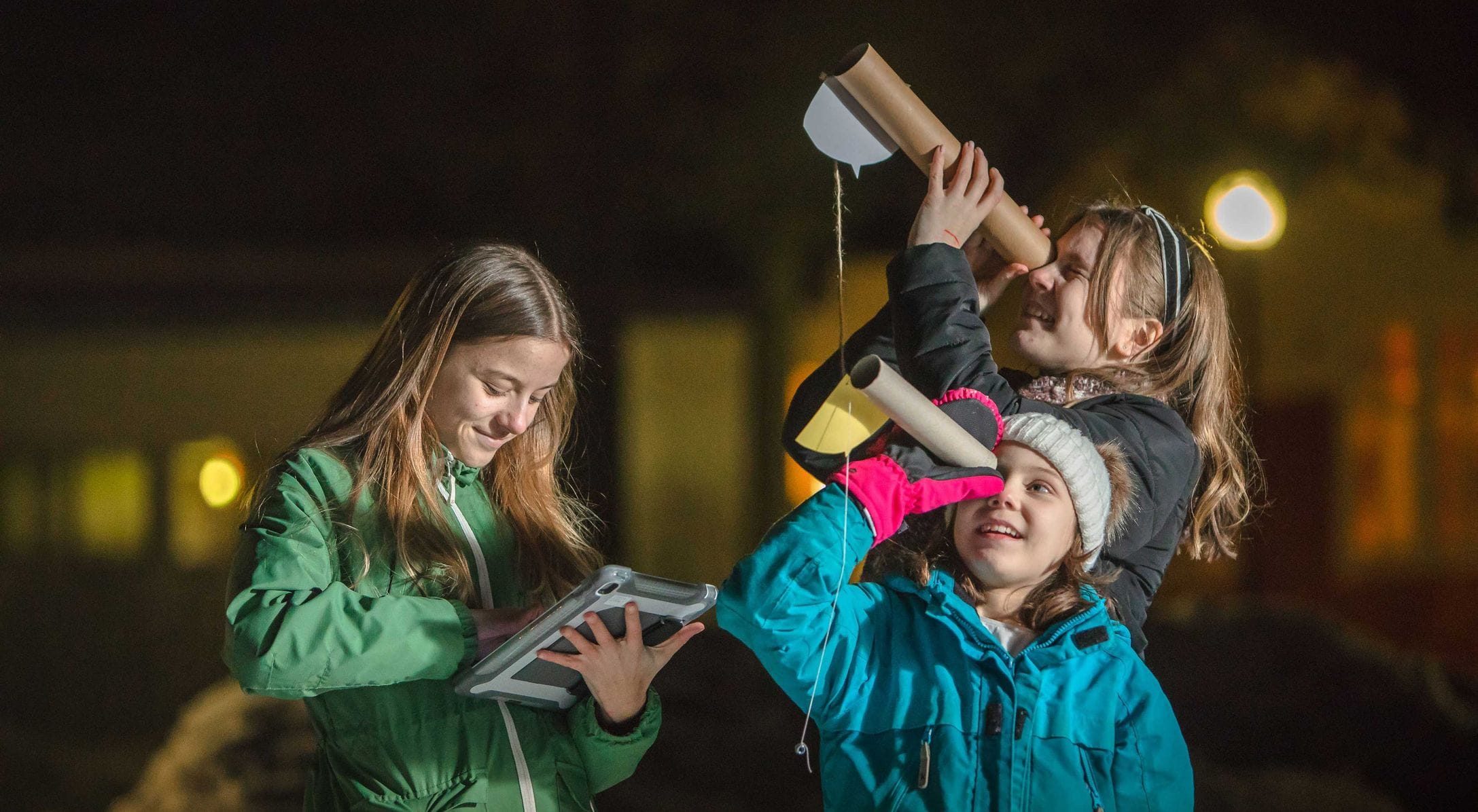 Three children explore the night sky. One holds a tablet while the other two look through simple telescopes made from tubes.